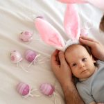 Man wearing funny bunny ears to his newborn son lying on bed among pink eggs. Celebrating Easter