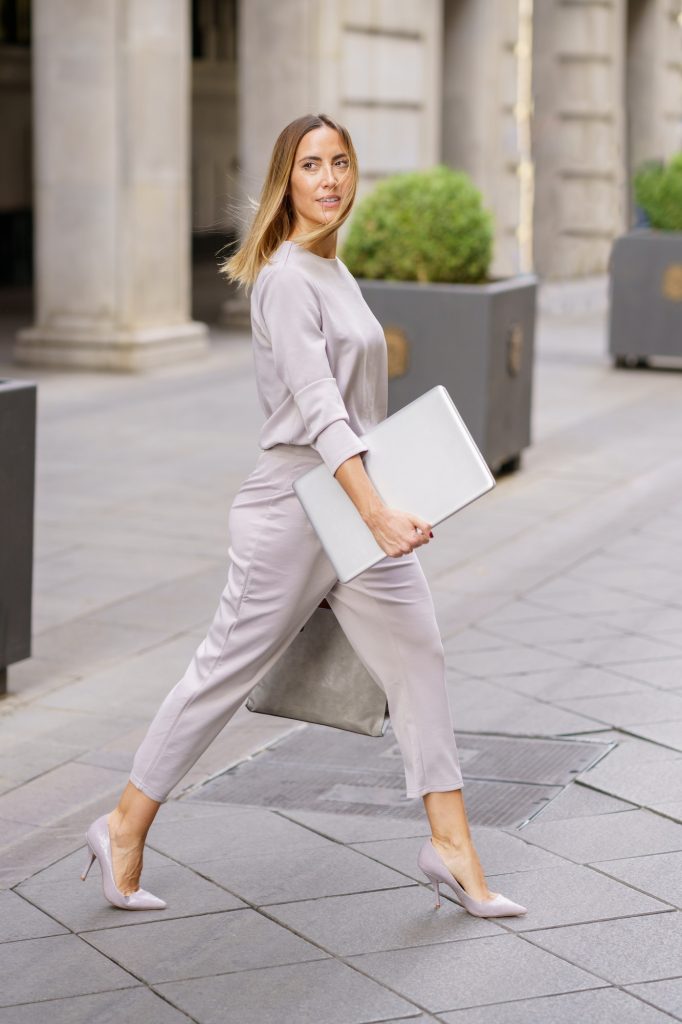 Self assured female manager with laptop crossing street near building